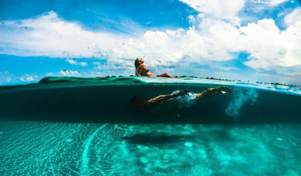 A stunning split-level photo shows a woman relaxing on a poolside deck under a bright blue sky, while underwater, a man swims effortlessly in crystal-clear turquoise water, capturing the vibrant essence of a sunny day.