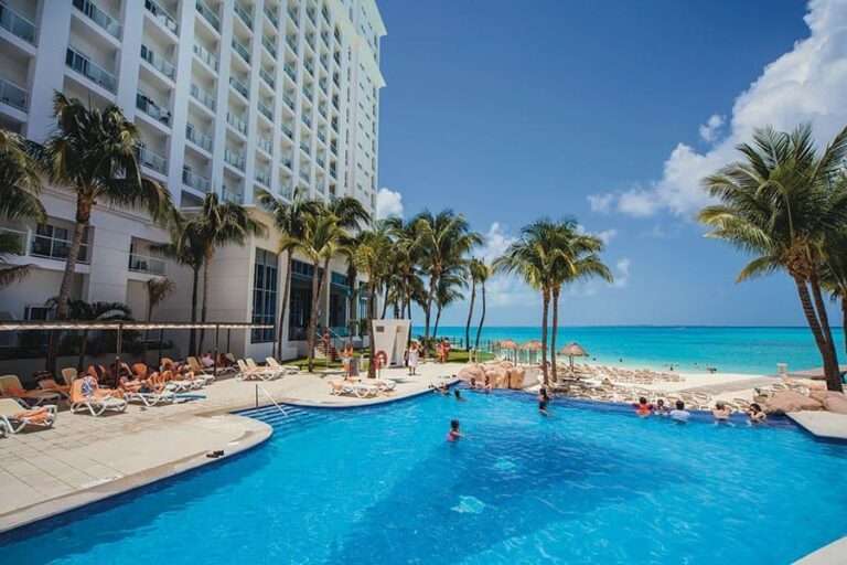 A tropical resort scene at the Riu Cancun Hotel features a large swimming pool surrounded by lounge chairs and palm trees. Guests relax by the pool, basking in the sunny weather. A beach with turquoise water stretches out in the background under a clear blue sky.