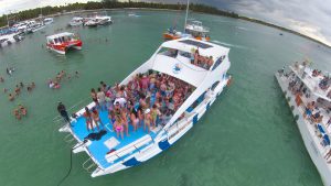 A large white boat crowded with people in swimwear floats on a greenish-blue ocean. Several other boats are nearby, and many people are swimming in the water. The background shows a cloudy sky and a distant treeline.