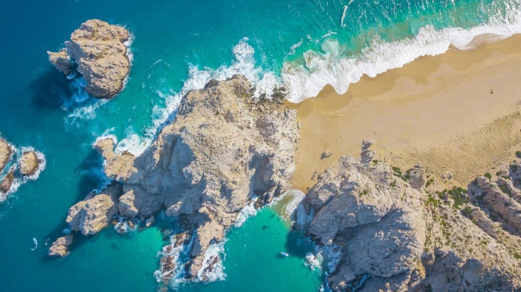 Aerial view of a rocky coastline with turquoise blue water. Waves crash against the rocks and a sandy beach, creating white foam, reminding one of when spring break adventures begin. The land extends into a rugged point surrounded by a sparkling ocean. Cabo San Lucas, B.C.S., Mexico