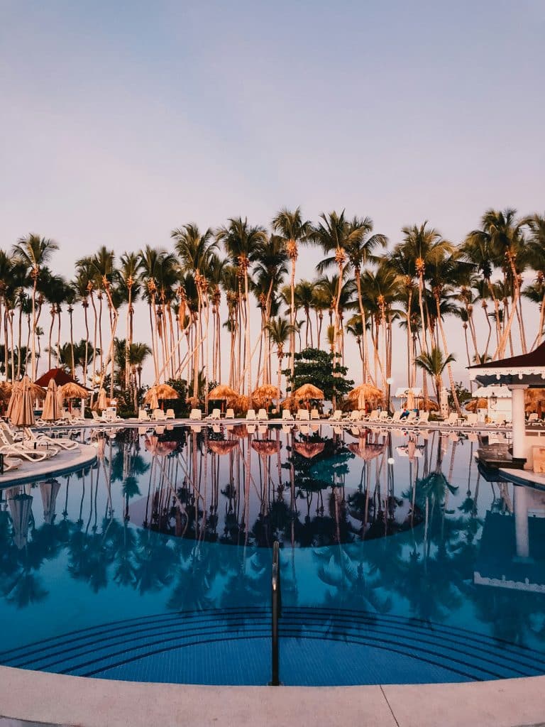 A luxurious outdoor pool at one of the best all-inclusive resorts in Mexico reflects tall palm trees and umbrellas under a clear sky. Lounge chairs surround the pool, creating a tranquil resort atmosphere perfect for a relaxing college spring break.