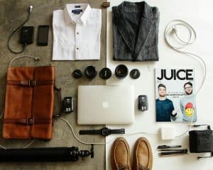 Flat lay of travel essentials including a white shirt, denim jacket, leather bag, camera lenses, a phone, laptop, magazine, chargers, tripod, watch, shoes, and pens on a gray surface.