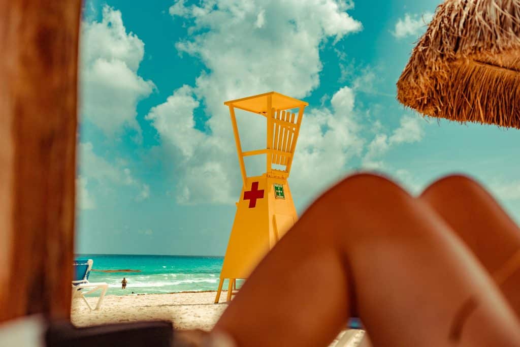 A beach scene with a yellow lifeguard tower featuring a red cross. The ocean is in the background under a partly cloudy sky. In the foreground, blurred legs of a sunbather are partially visible, and a straw thatched umbrella frames the scene.