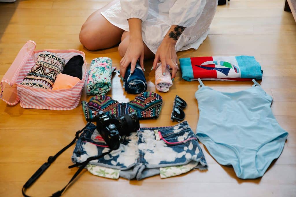 A person sits on the floor organizing travel items: a pink-striped box with clothes, rolled garments, a blue swimsuit, patterned shorts, a colorful top, sunglasses, and a camera. The person wears a white outfit, and their hands are arranging the items.