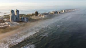 Aerial view of a coastal city with tall buildings along a foggy shoreline. Waves gently crash onto the beach, and the horizon stretches into the distance under a clear sky.