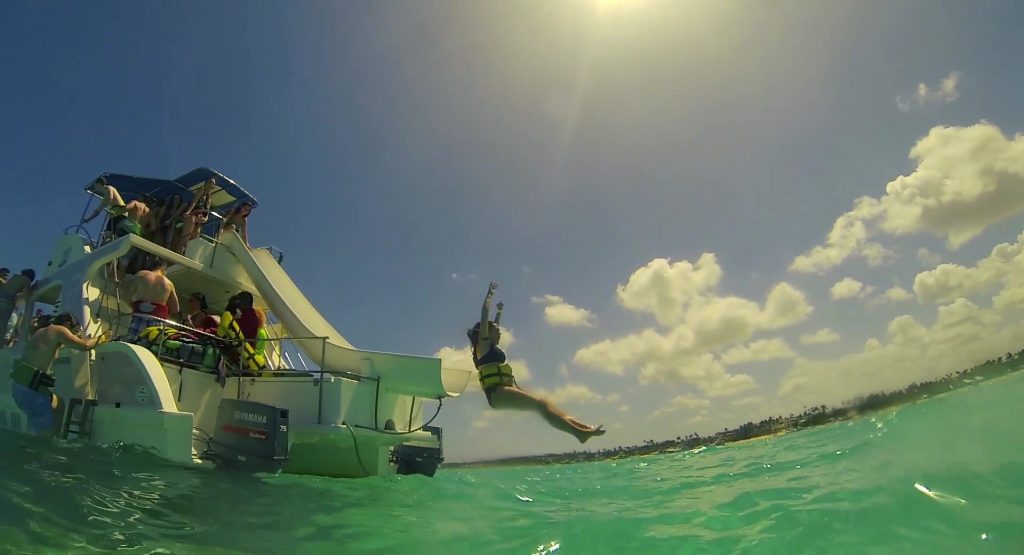 A person in a life jacket jumps off a boat slide into clear turquoise water. Other people watch from the deck. The sky is bright and sunny with scattered clouds.