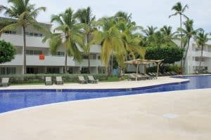 A tropical resort features a large, curved pool surrounded by palm trees. Several lounge chairs and shaded spots line the poolside near a modern, white multi-story building. A bright orange towel hangs from a balcony.