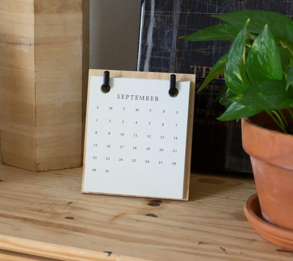 A desk calendar displaying the month of September is placed on a wooden surface. Next to it, there is a green potted plant and a partially visible book with a black cover.