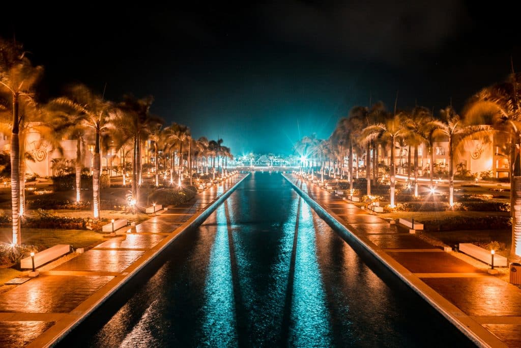 A nighttime scene shows a long, illuminated pathway lined with palm trees and benches, bordered by a reflective water feature. Warm lights enhance the serene ambiance, creating a symmetrical view towards a bright focal point in the distance at the Hard Rock Hotel & Casino Punta Cana, Punta Cana, Dominican Republic