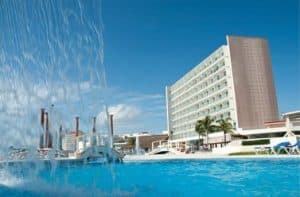 View of a hotel with a tall modern building, a pool in the foreground featuring cascading water, and several lounge chairs. The sky is bright and clear, with a few scattered clouds. Palm trees are visible by the poolside.