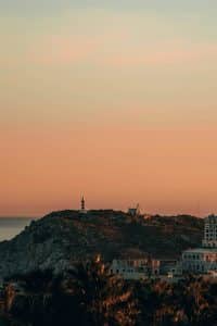 Sunset view of a hilly landscape with a lighthouse on top of a hill, surrounded by scattered buildings and lush vegetation. The sky is painted with warm orange and pink hues, adding a tranquil atmosphere to the scene. Cabo San Lucas, BCS, Mexico