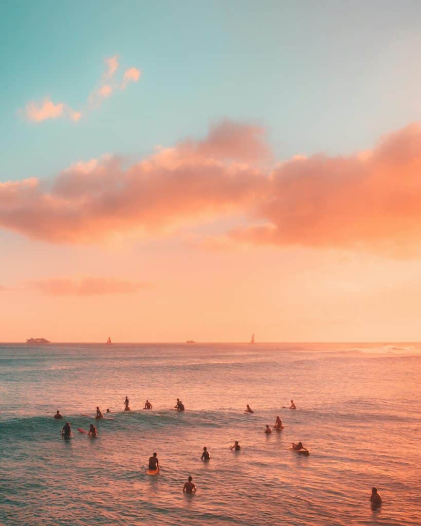 A group of surfers sit on their boards in calm ocean waters under a vibrant sunset sky. The sky is streaked with orange and pink hues, and a few sailboats are visible in the distance.