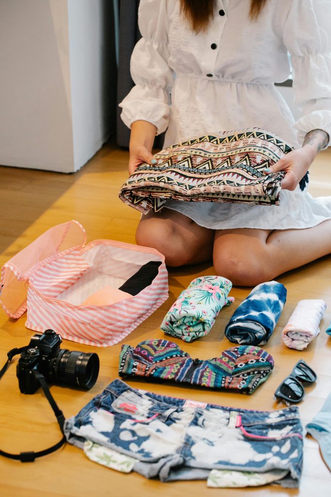 A person in a white dress is packing a suitcase on the floor. Items include a folded patterned fabric, rolled clothes, a camera, sunglasses, a floral pouch, and denim shorts.