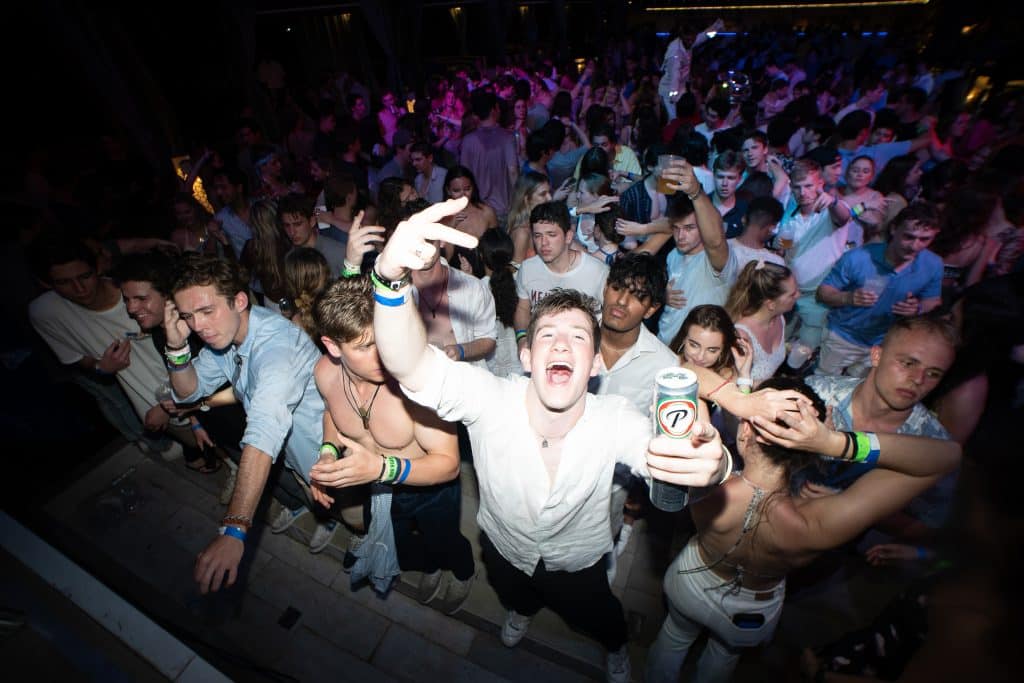 A crowded party scene with people dancing and enjoying themselves. In the foreground, a person in a white shirt holds up a can and gestures energetically towards the camera. The background is filled with colorful lights and a lively atmosphere.