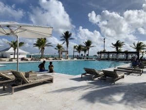 A tranquil resort swimming pool with sun loungers and parasols. Two people sit at the pool’s edge, with palm trees and a cloudy sky in the background. The ocean can be seen beyond the pool area.