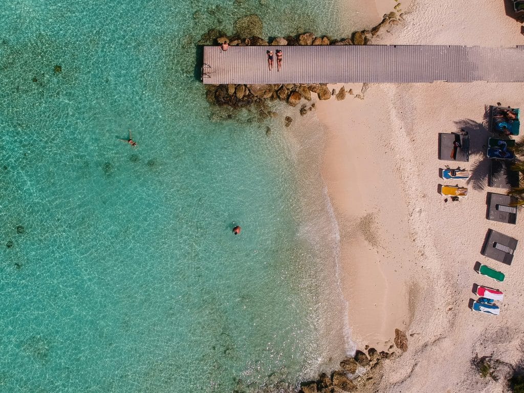 Aerial view of a tranquil beach scene with clear turquoise water. A wooden pier extends over the water, where two people relax. Sunbeds and umbrellas are scattered on the sandy shore, and a few swimmers enjoy the pristine sea.