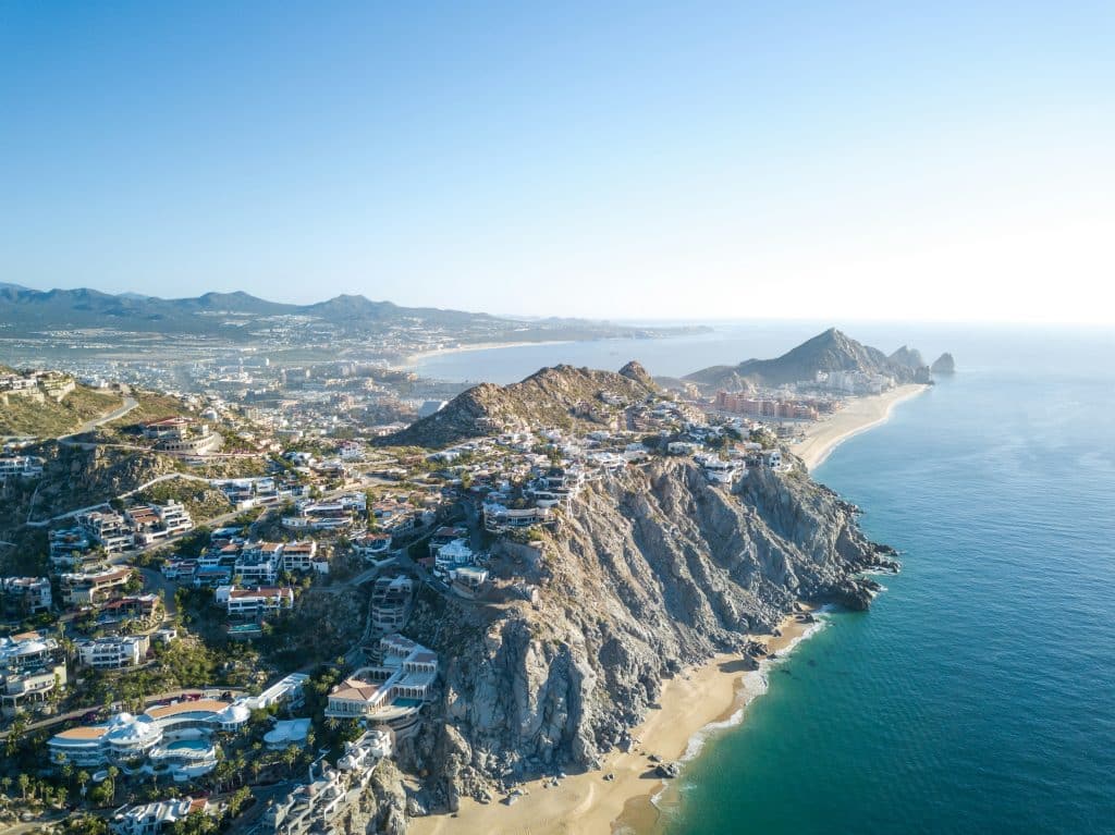 Aerial view of a coastal landscape with rocky cliffs and sandy beaches. Luxurious houses are perched on the cliffs, overlooking the clear blue ocean. The horizon is visible under a clear blue sky.