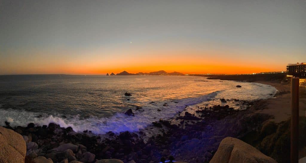 A stunning coastal sunset with a vibrant orange and blue sky. The ocean waves gently crash against a rocky shoreline. Distant hills are silhouetted by the sunset, and a small crescent moon is visible in the sky.