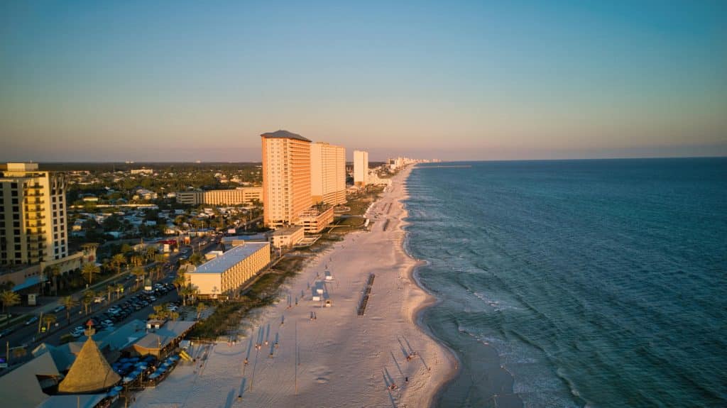 Aerial view of a sandy beach with high-rise buildings along the shoreline. The beach curves alongside the calm blue ocean under a clear sky. Umbrellas and beachgoers are scattered along the sand.