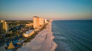 Aerial view of a sandy beach with high-rise buildings along the shoreline. The beach curves alongside the calm blue ocean under a clear sky. Umbrellas and beachgoers are scattered along the sand.