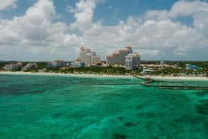 Aerial view of a coastal resort with multiple tall buildings surrounded by lush greenery. The turquoise ocean and sandy beach are in the foreground, with a mix of white clouds and blue sky above.