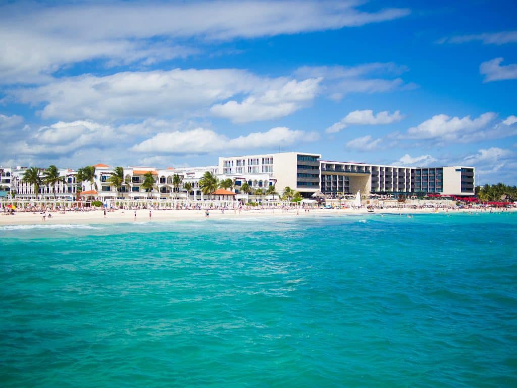 Scenic view of a beachfront resort with modern buildings along a sandy shore. The turquoise ocean is in the foreground, and a clear blue sky with scattered clouds is in the background. Palm trees dot the landscape.
