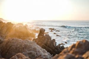 Rocky coastline with the sun setting over the ocean, casting a warm glow on the rocks and waves. The sea appears calm with gentle waves washing against the shore. Cabo San Lucas, Mexico