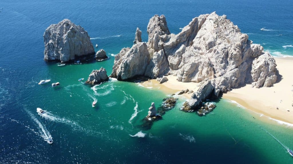 Aerial view of a picturesque coastal scene with jagged rock formations rising from turquoise waters near a sandy beach. Several boats are seen navigating around the formations, leaving white trails behind. The ocean extends into the distance.