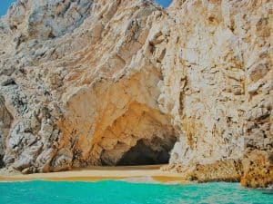 A rocky cave opening along a sandy beach with turquoise water in the foreground. The large, jagged rock formations create a dramatic entrance to the cave, and the clear blue sky is visible above.