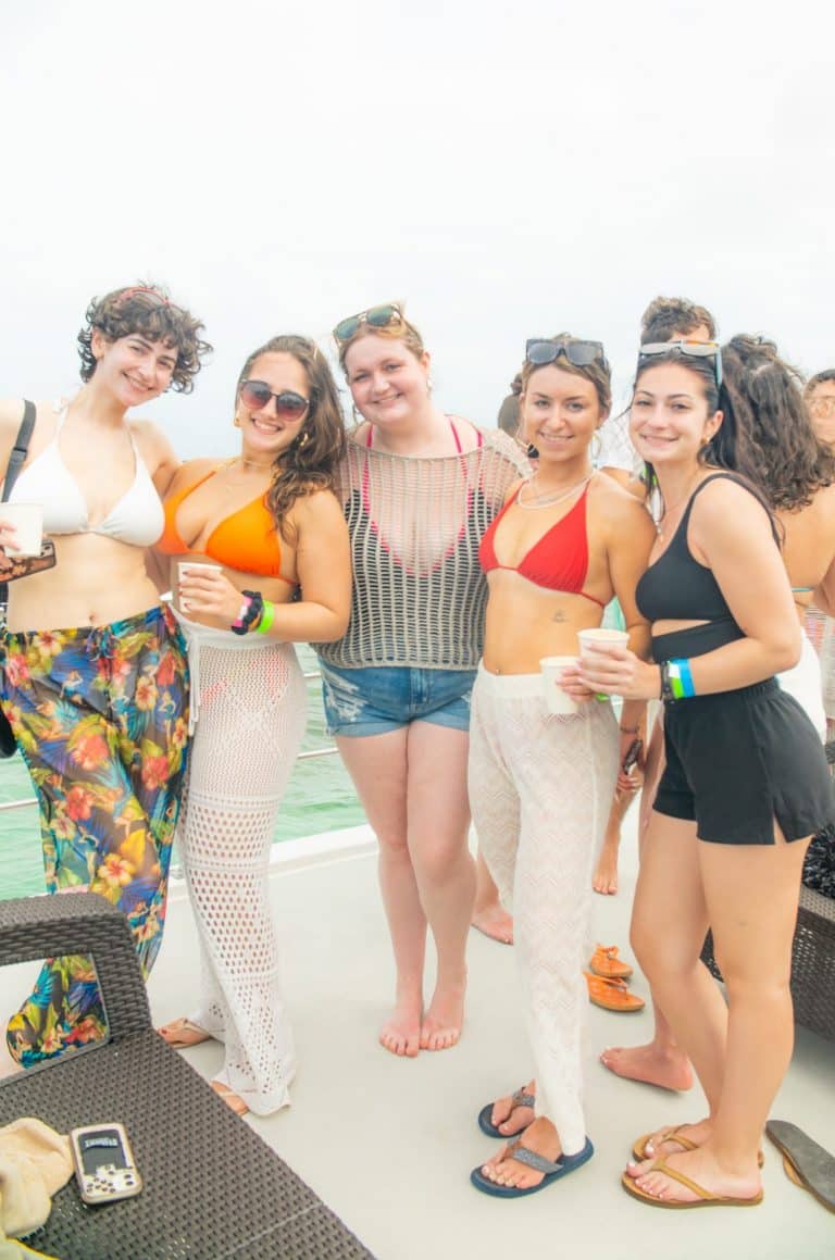 A group of five people poses together on a boat deck. They are dressed in swimsuits and beach attire. Four of them hold drinks, and they are all smiling at the camera. The background shows a cloudy sky and the sea.