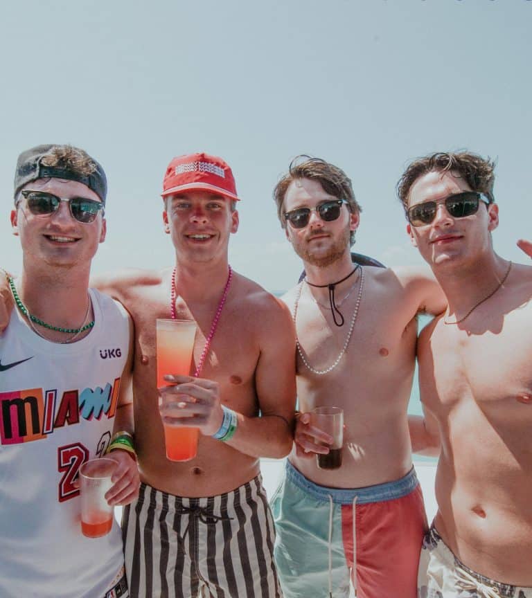 Four men stand together on a sunny beach. They are shirtless, wearing sunglasses, and holding drinks. One wears a Miami jersey, another a red cap. All are smiling and seem to be enjoying a lively atmosphere.