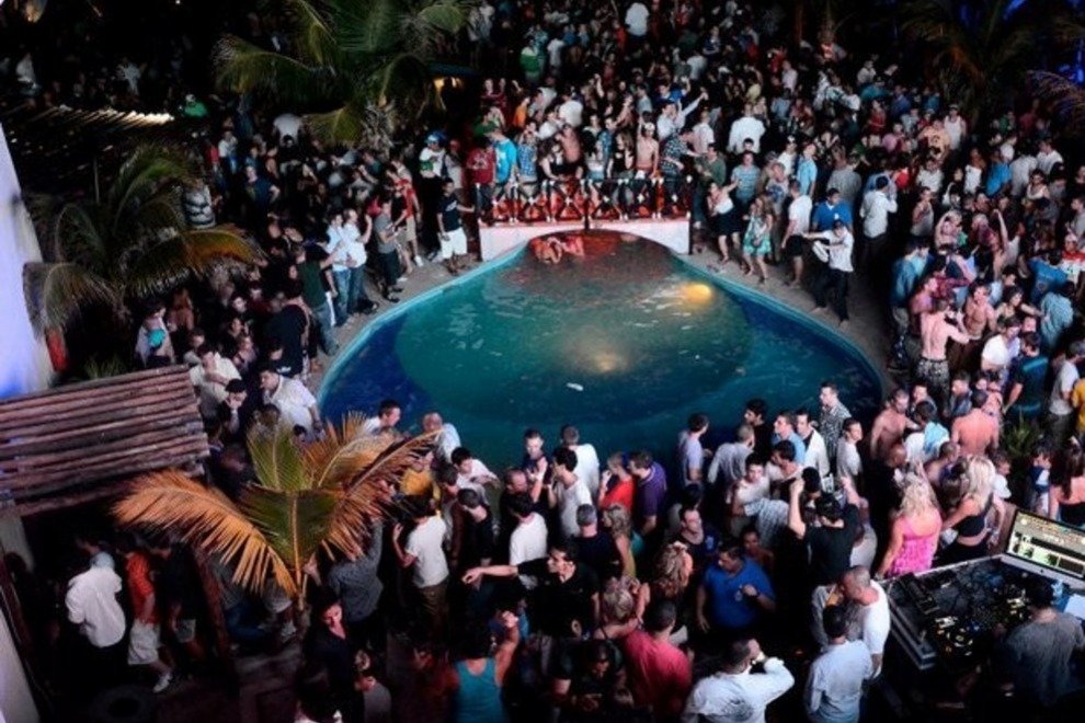 A large crowd of people gathers around a small pool at a lively outdoor event. Some attendees are standing near palm trees, and others are around a DJ booth. Colorful lights illuminate the scene, creating a festive atmosphere.