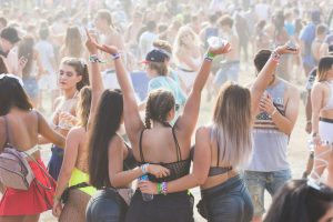 A group of people enjoying an outdoor festival. Three women with their backs to the camera raise their arms in celebration. The crowd around them is lively, with people dancing and socializing in a sunlit, dusty atmosphere.