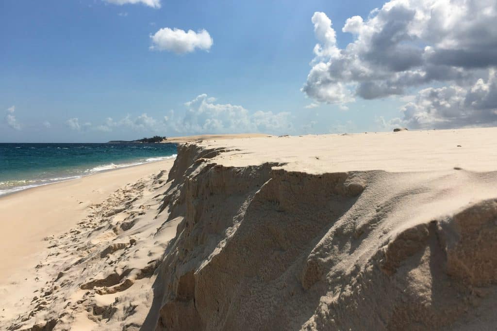 A sandy beach with a sharp sand dune edge overlooking the ocean under a bright blue sky with scattered clouds. Waves gently lap the shore, and footprints are visible in the sand.