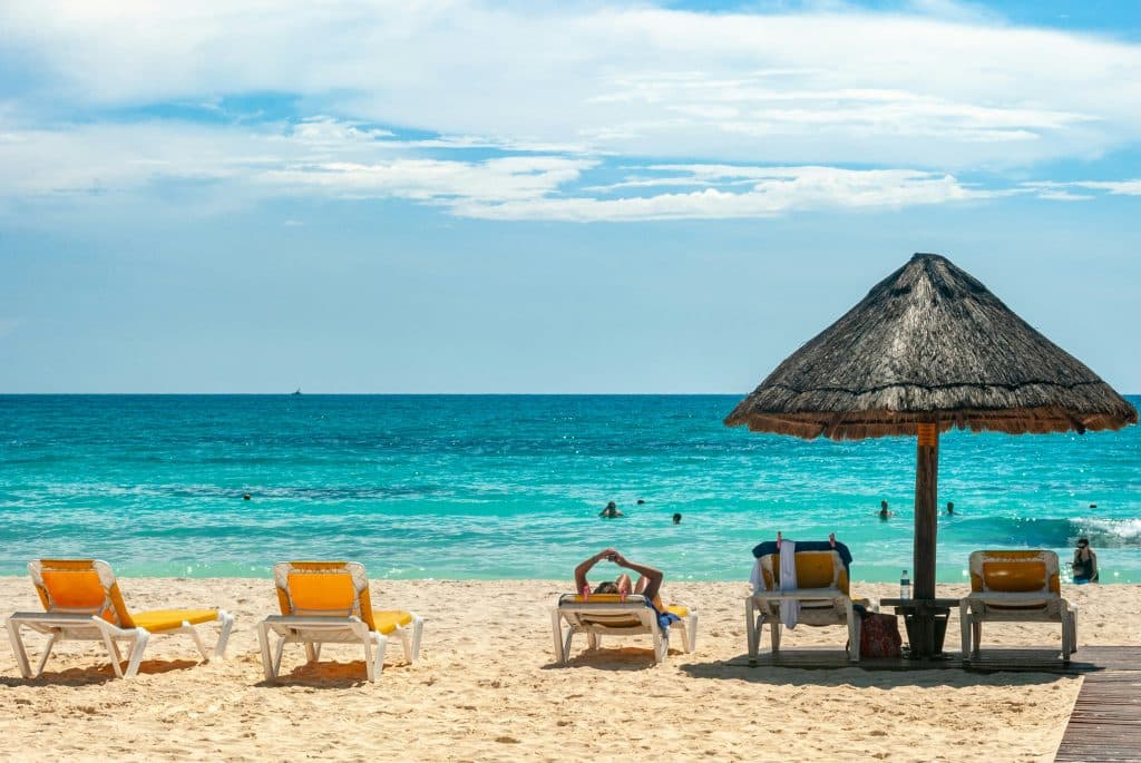 Sandy beach with yellow lounge chairs and a thatched umbrella facing turquoise ocean; people are swimming and relaxing under a partly cloudy sky.