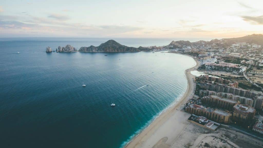 Aerial view of a coastal city with sandy beaches, blue ocean, boats on the water, and rocky hills in the distance at sunset. Buildings line the shore on the right side of the image.