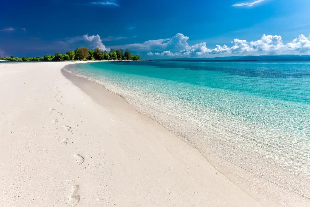 A pristine white sand beach with clear turquoise water, gentle waves, and footprints leading along the shoreline; lush green trees and a blue sky with scattered clouds in the background.
