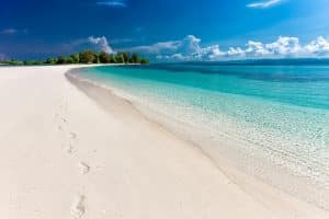 A pristine white sand beach with clear turquoise water, gentle waves, and footprints leading along the shoreline; lush green trees and a blue sky with scattered clouds in the background.