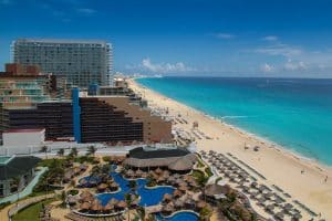 Aerial view of beachfront resorts in Cancun, Mexico, with modern hotels, pools, palm trees, white sand beach, and turquoise ocean under a bright blue sky. Rows of beach chairs line the shore.