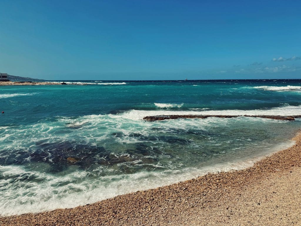 A clear, sunny day at a rocky beach with turquoise waves gently crashing onto the shore; the horizon is visible under a blue sky with a few scattered clouds.