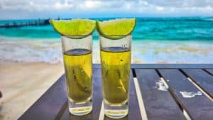 Two shot glasses filled with golden liquid and topped with lime wedges sit on a table, with a sandy beach and turquoise ocean waves in the background.