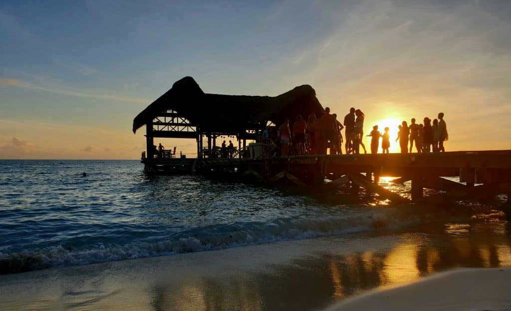 Silhouetted people gather on a pier with a thatched-roof hut, watching the sunset over calm ocean waves; golden sunlight reflects on wet sand and water.