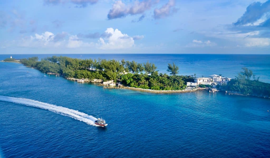 A small motorboat speeds across clear blue water near a lush, green island with a few white buildings, under a partly cloudy sky.