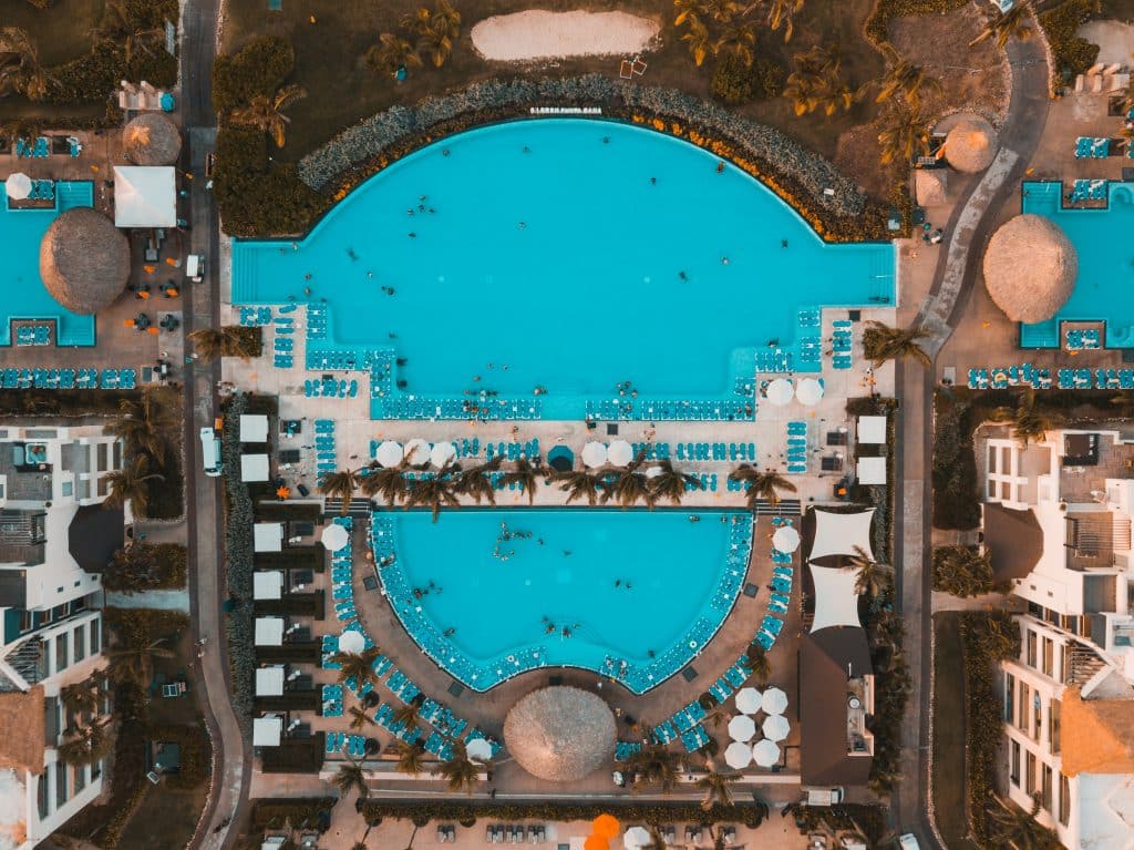 Aerial view of a large, rectangular swimming pool surrounded by palm trees, lounge chairs, and resort buildings. People are swimming and relaxing poolside, creating a vibrant, tropical atmosphere.