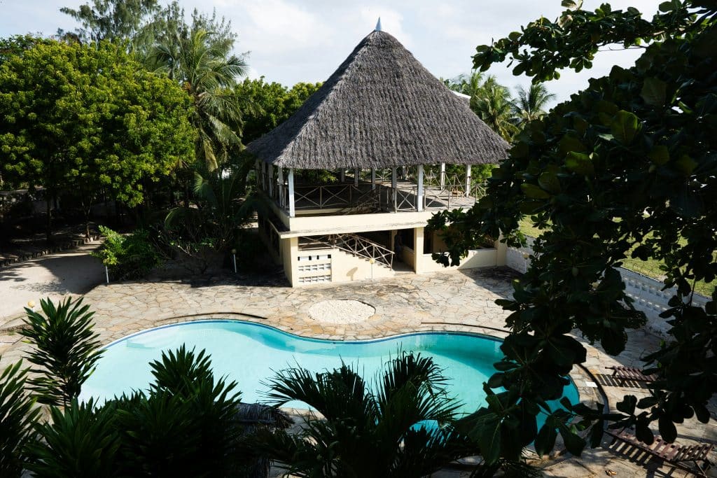 A tropical outdoor swimming pool with curved edges is surrounded by stone decking, lush green trees, and a thatched-roof pavilion on stilts, creating a relaxing vacation setting.