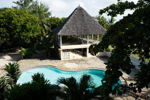 A tropical outdoor swimming pool with curved edges is surrounded by stone decking, lush green trees, and a thatched-roof pavilion on stilts, creating a relaxing vacation setting.
