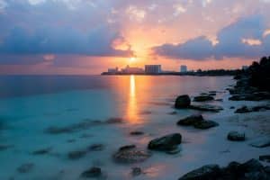 Sunset over a calm beach with gentle waves, rocks in the foreground, and city buildings silhouetted against a colorful sky with glowing clouds.