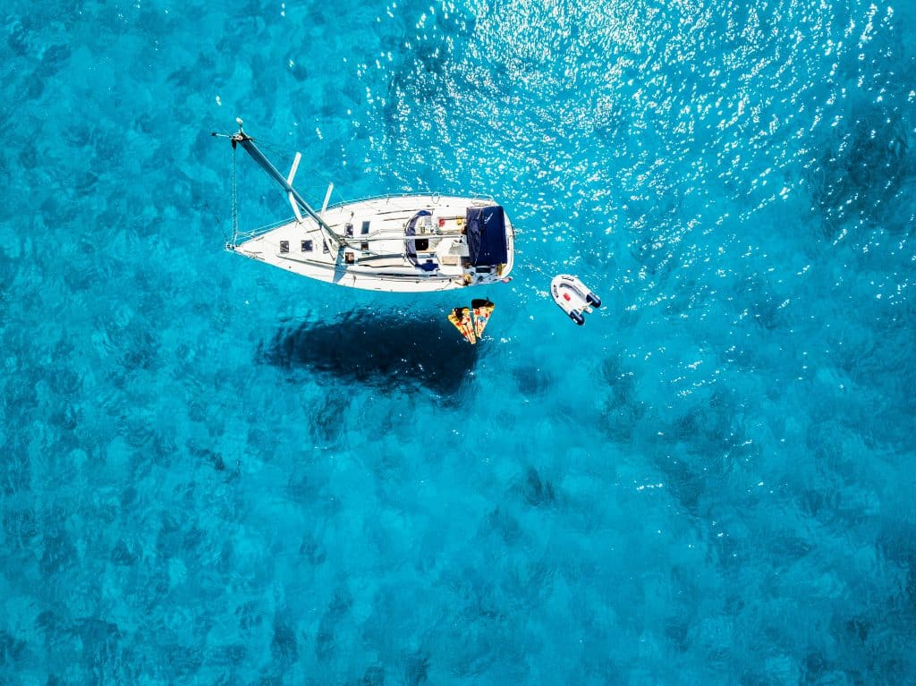 Aerial view of a white sailboat anchored in clear blue water with two people relaxing on orange floats beside it and a small inflatable boat nearby. Sunlight sparkles on the surface of the sea.