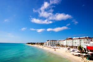 Modern beachfront hotels line a sandy shore beside turquoise ocean water under a bright blue sky with scattered clouds; people relax and swim along the lively beach.