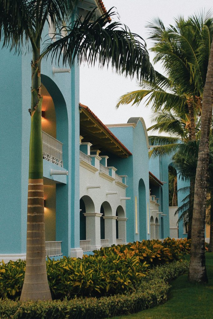 A pastel blue building with white balconies and arched doorways, surrounded by palm trees and lush green landscaping, under a clear sky.
